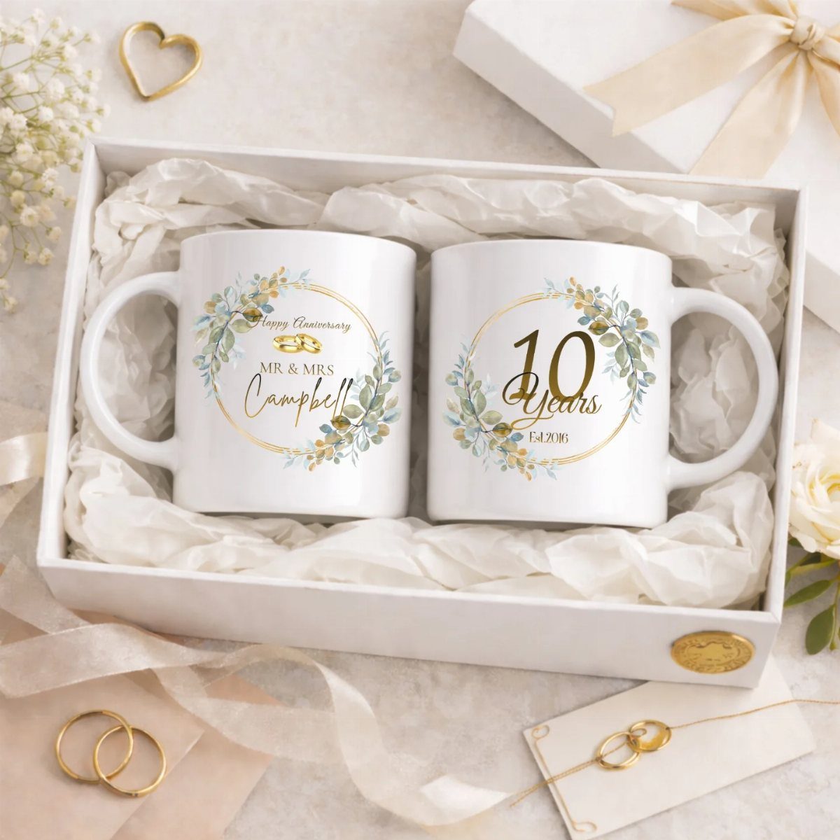 Two white anniversary coffee mugs with gold floral designs inside a gift box.
