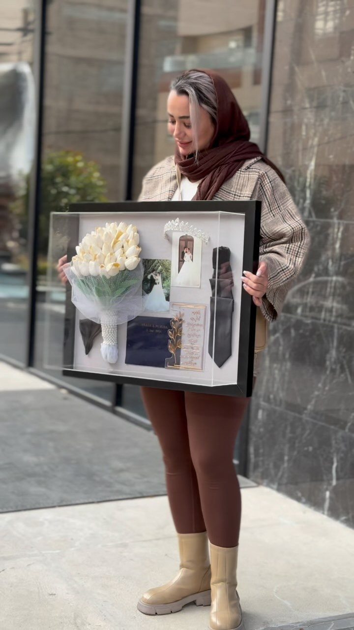 Woman holding a shadow box containing white flowers, wedding photos, and a black tie.