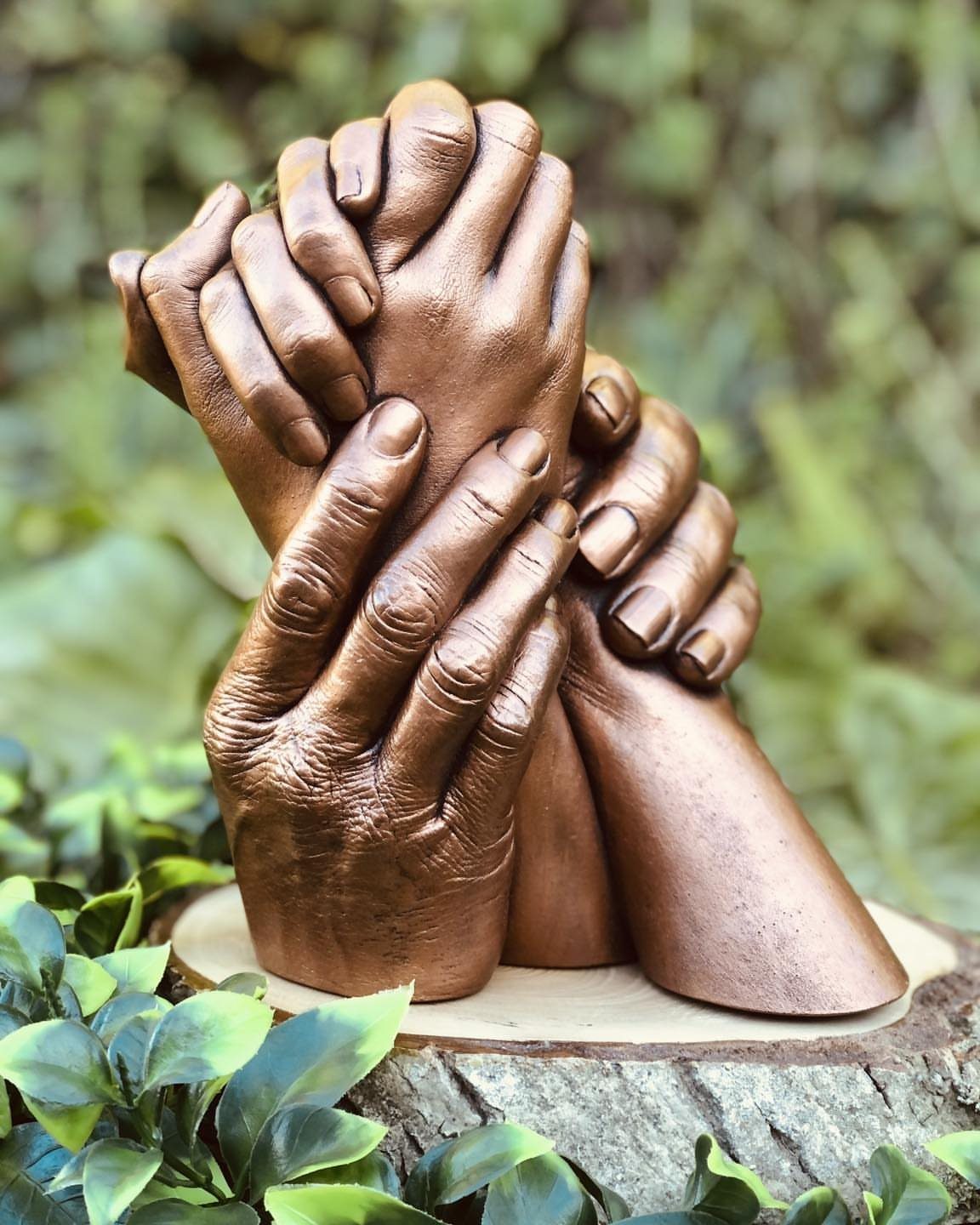 Bronze sculpture of intertwined hands resting on a wooden base amidst green leaves.