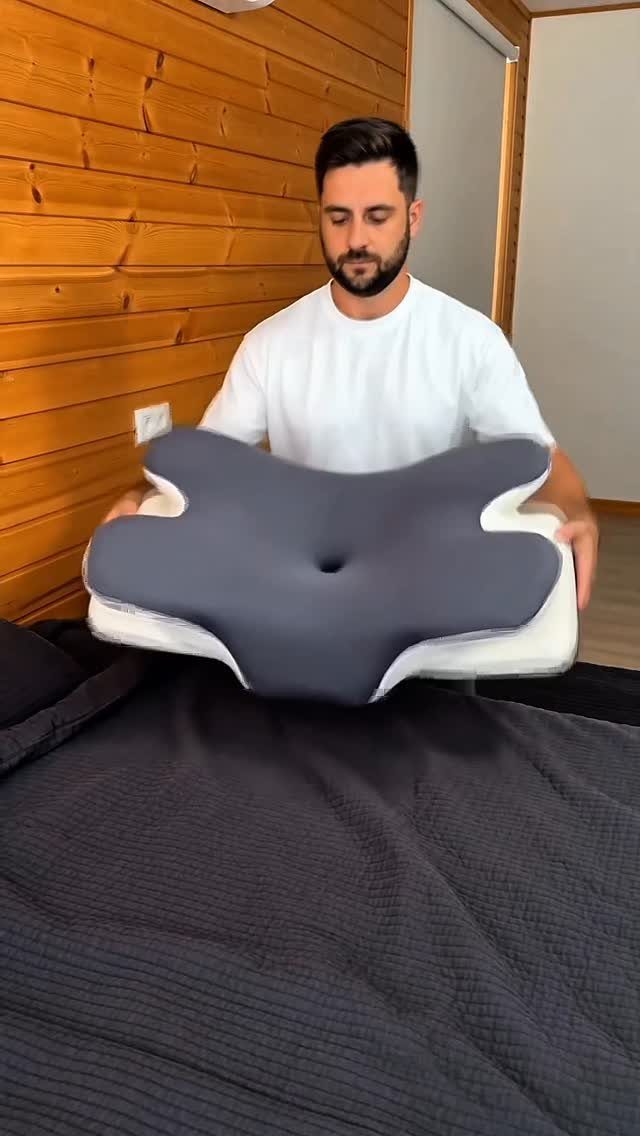 Man holding a grey ergonomic memory foam pillow over a dark grey bed
