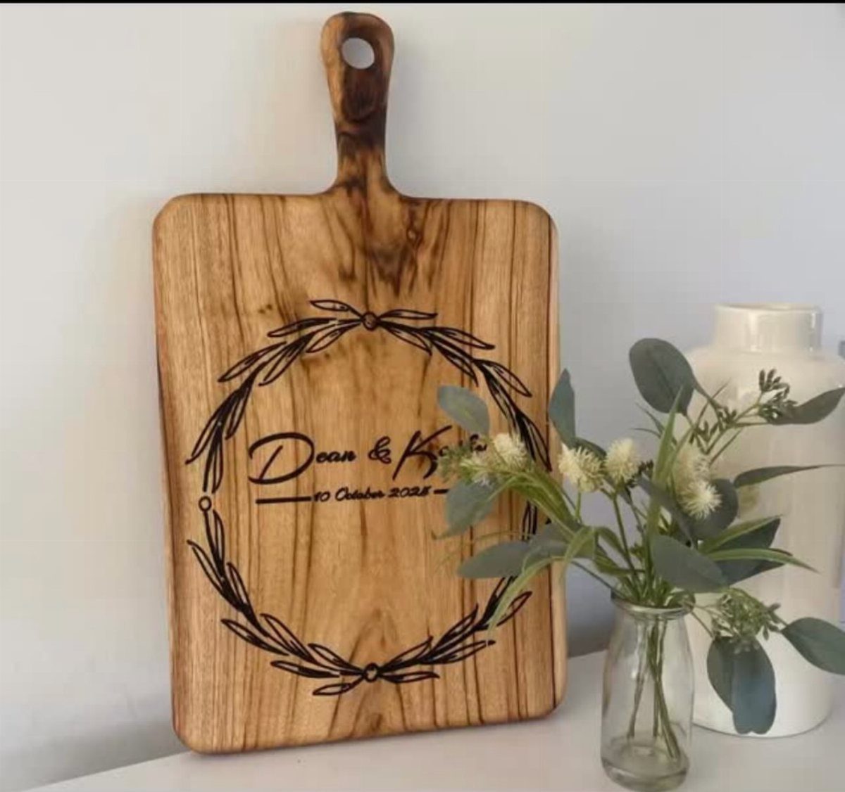 Engraved wooden cutting board with a wreath and names leaning against a white wall.