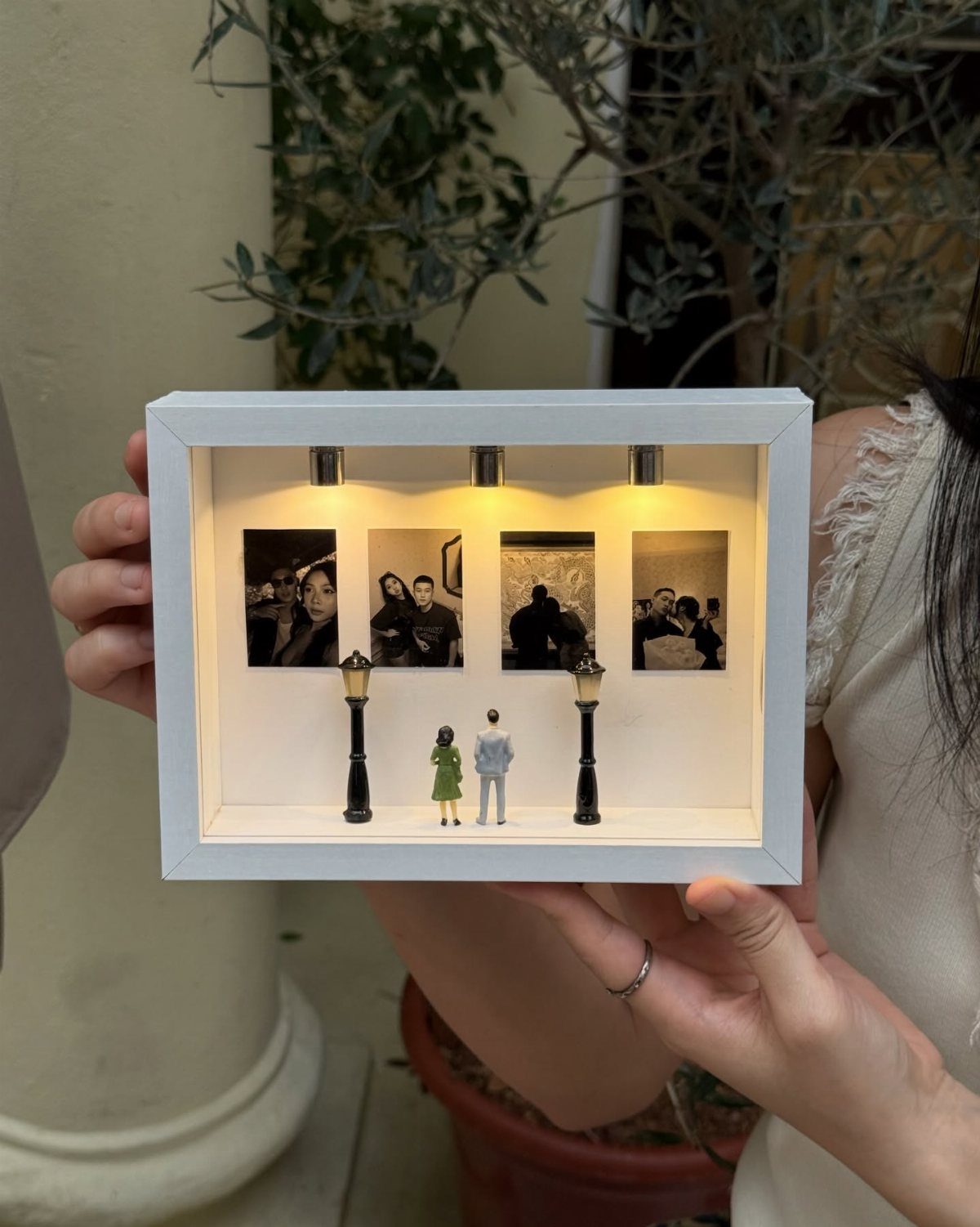 White 3D shadow box with miniature couple, street lamps, and small personal photographs.