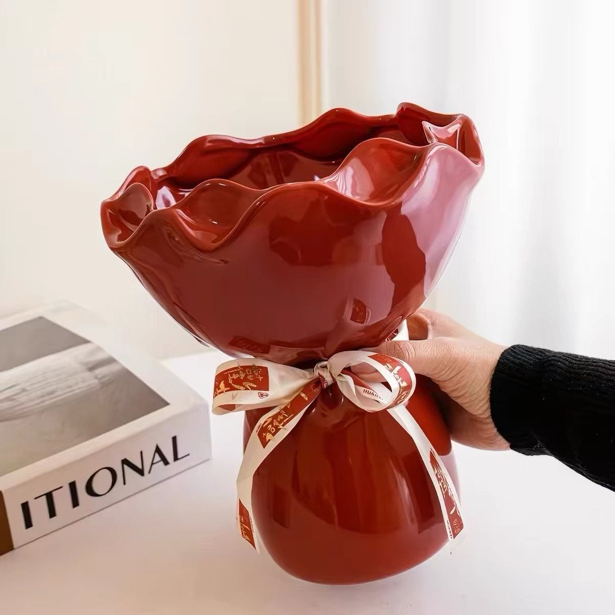Red ceramic vase with ruffled edges and a white ribbon held by a hand.
