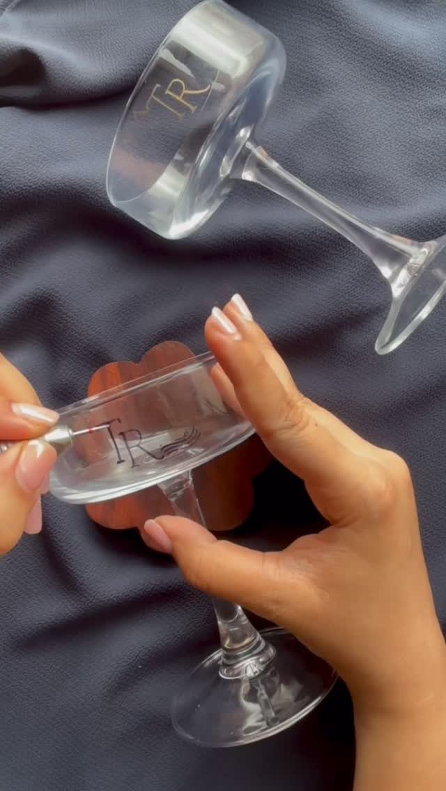 Hands engraving initials into a clear glass champagne flute on grey fabric.