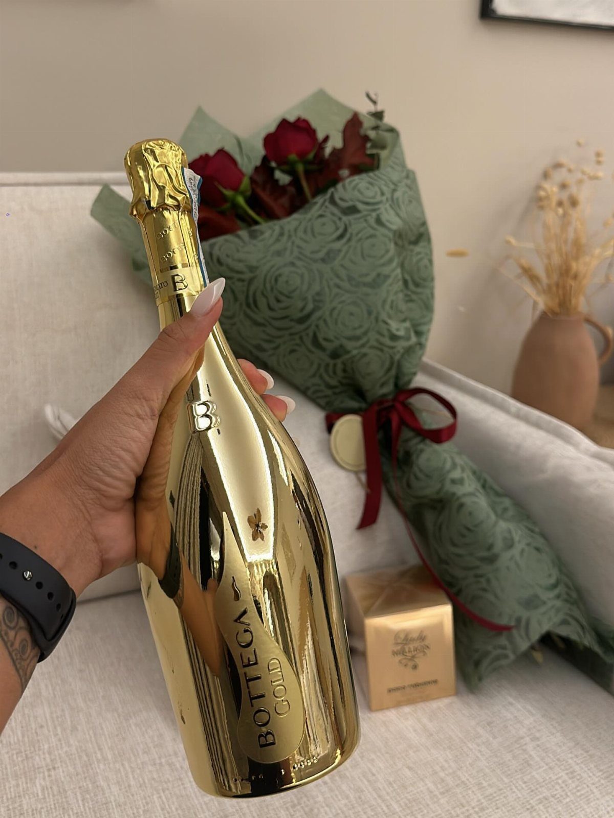 Gold champagne bottle held in hand with red roses and gift box behind.
