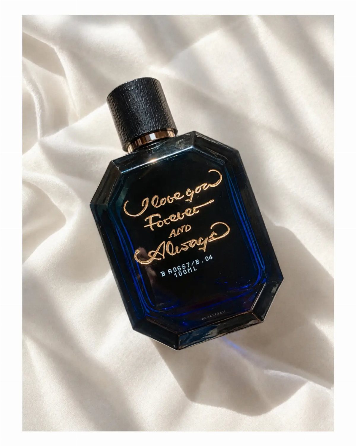 Dark blue octagonal perfume bottle with gold cursive text on white draped fabric