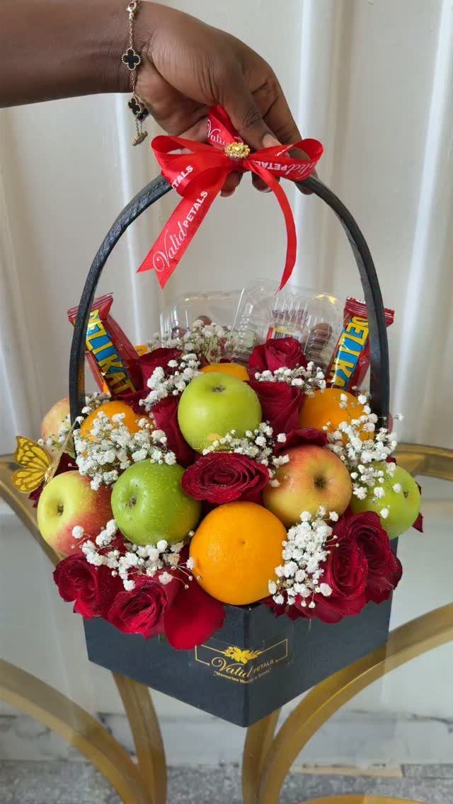 Gift basket containing red roses, green apples, and oranges with a red ribbon.