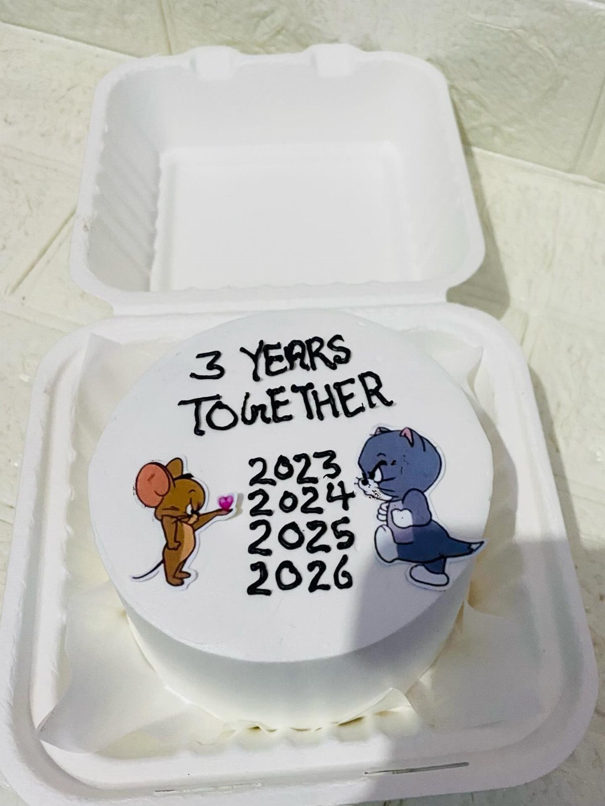 White frosted cake with Tom and Jerry cartoons and anniversary text in a container.