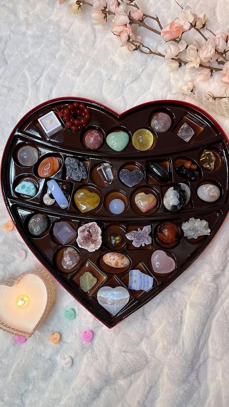 Heart-shaped box of colorful gemstones and crystals on white fabric with a candle.