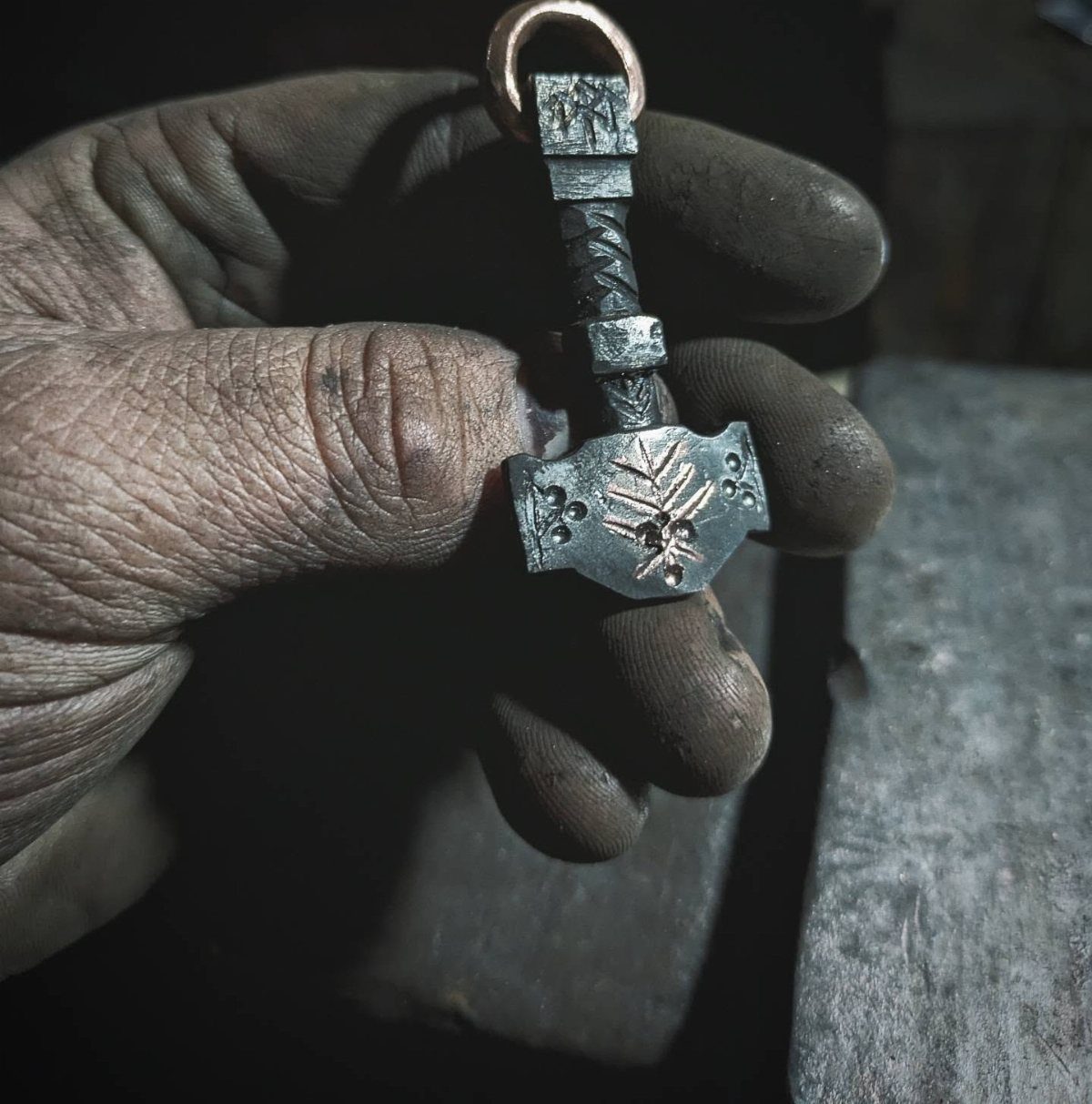 Handcrafted metal hammer pendant held by a soot-covered hand in a workshop
