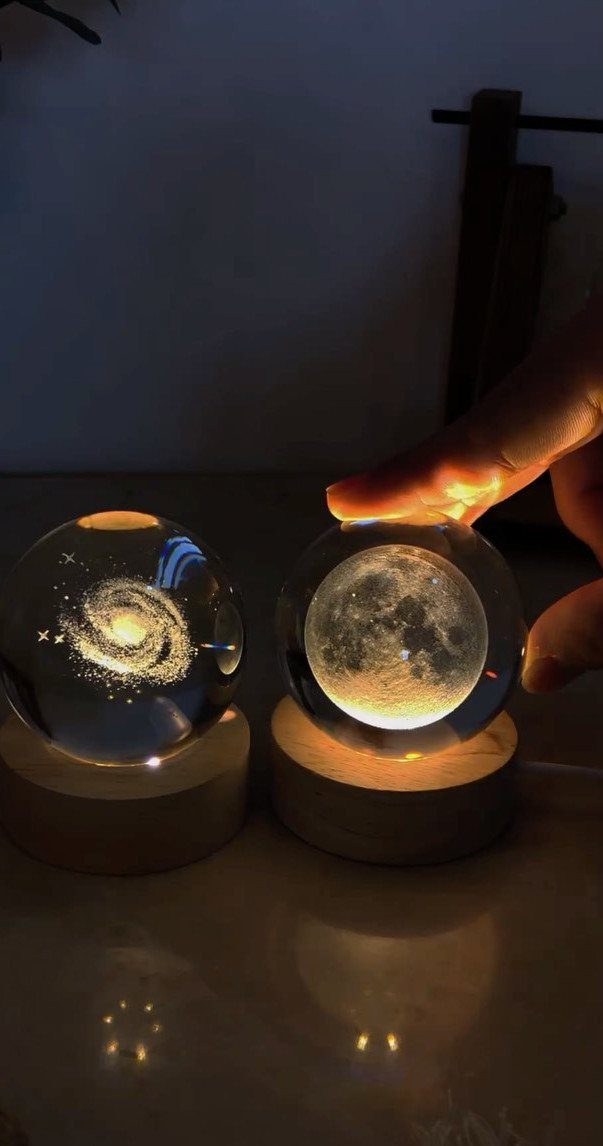 Two glowing glass spheres with galaxy and moon designs on wooden bases.
