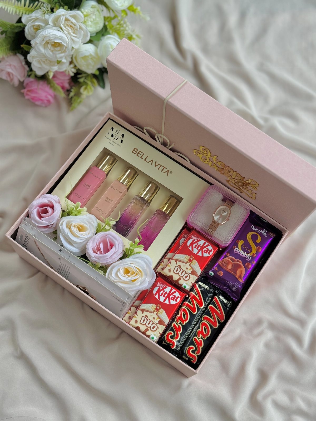 Pink gift box with perfumes, chocolates, a watch, and artificial roses on fabric.
