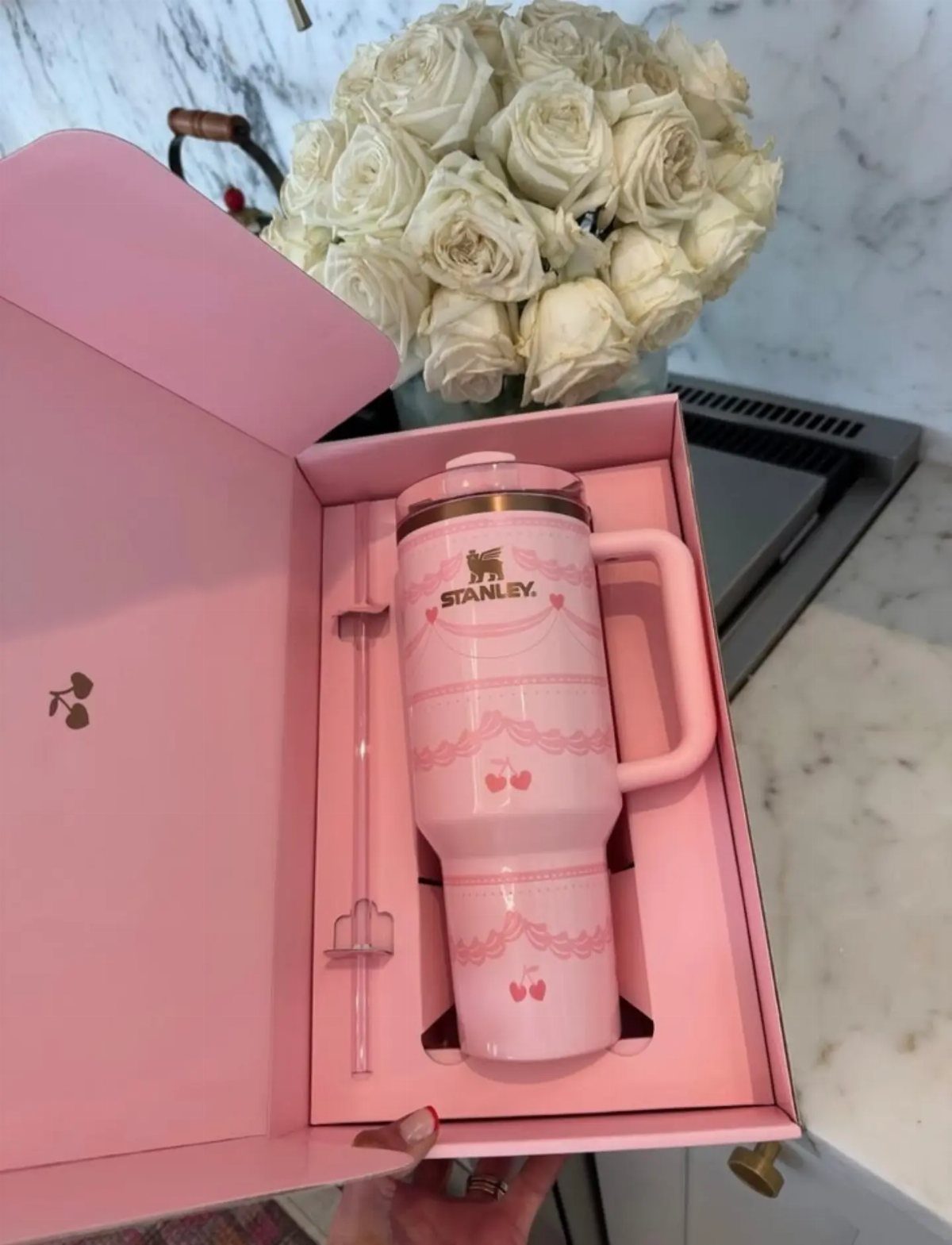Pink Stanley tumbler with cherry patterns in a pink gift box beside white roses.