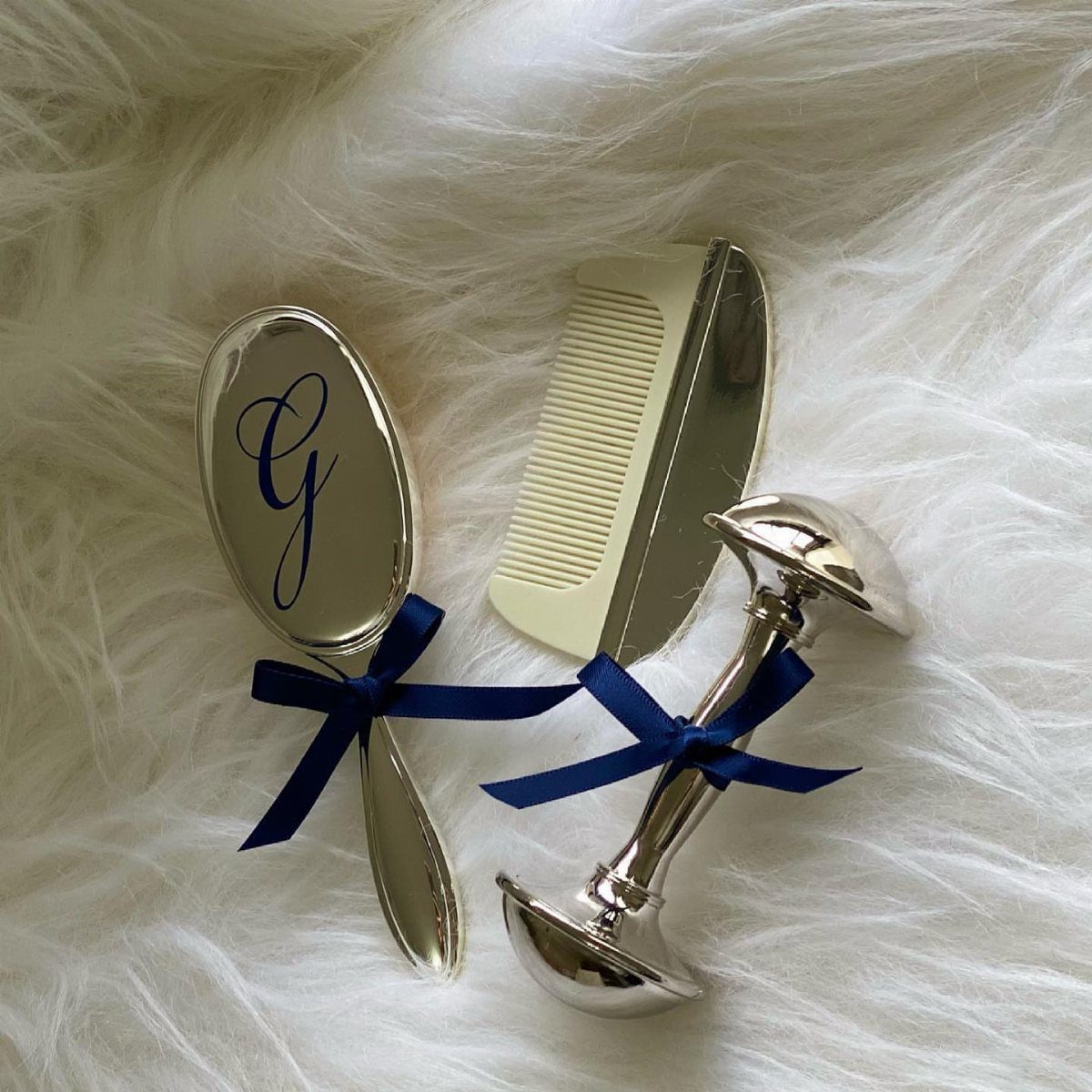 Silver baby brush, comb, and rattle with blue ribbons on white fur.