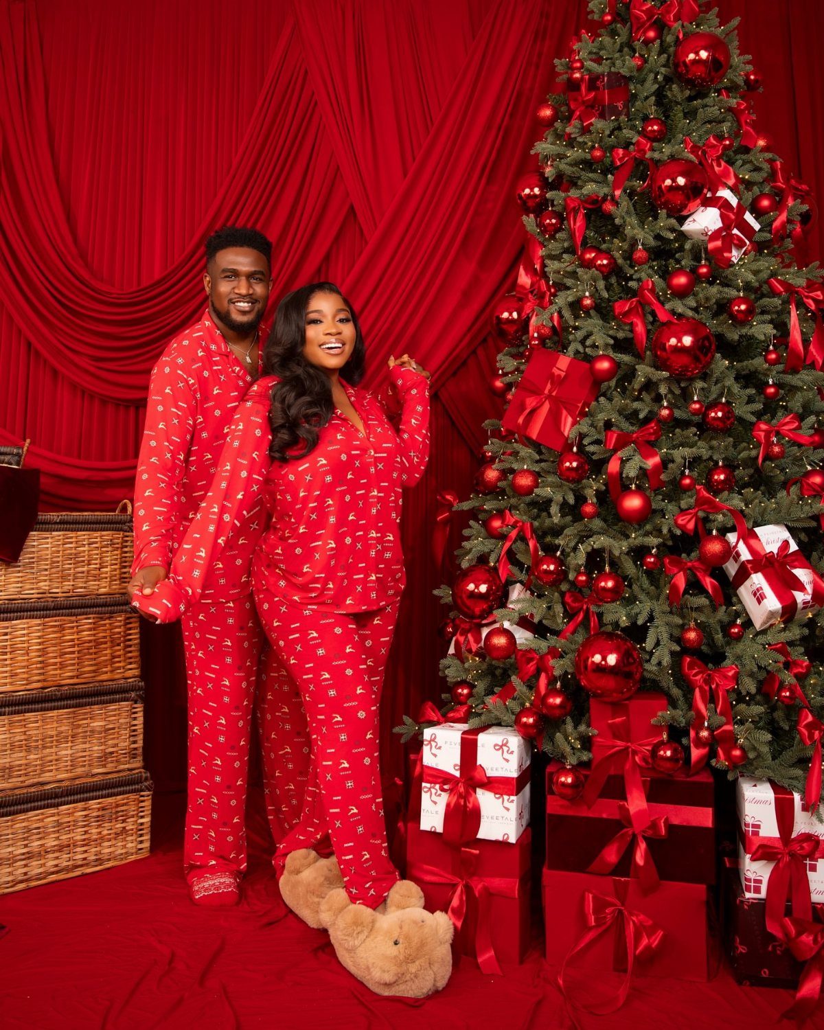 Couple in matching red pajamas standing by a decorated Christmas tree with gifts.