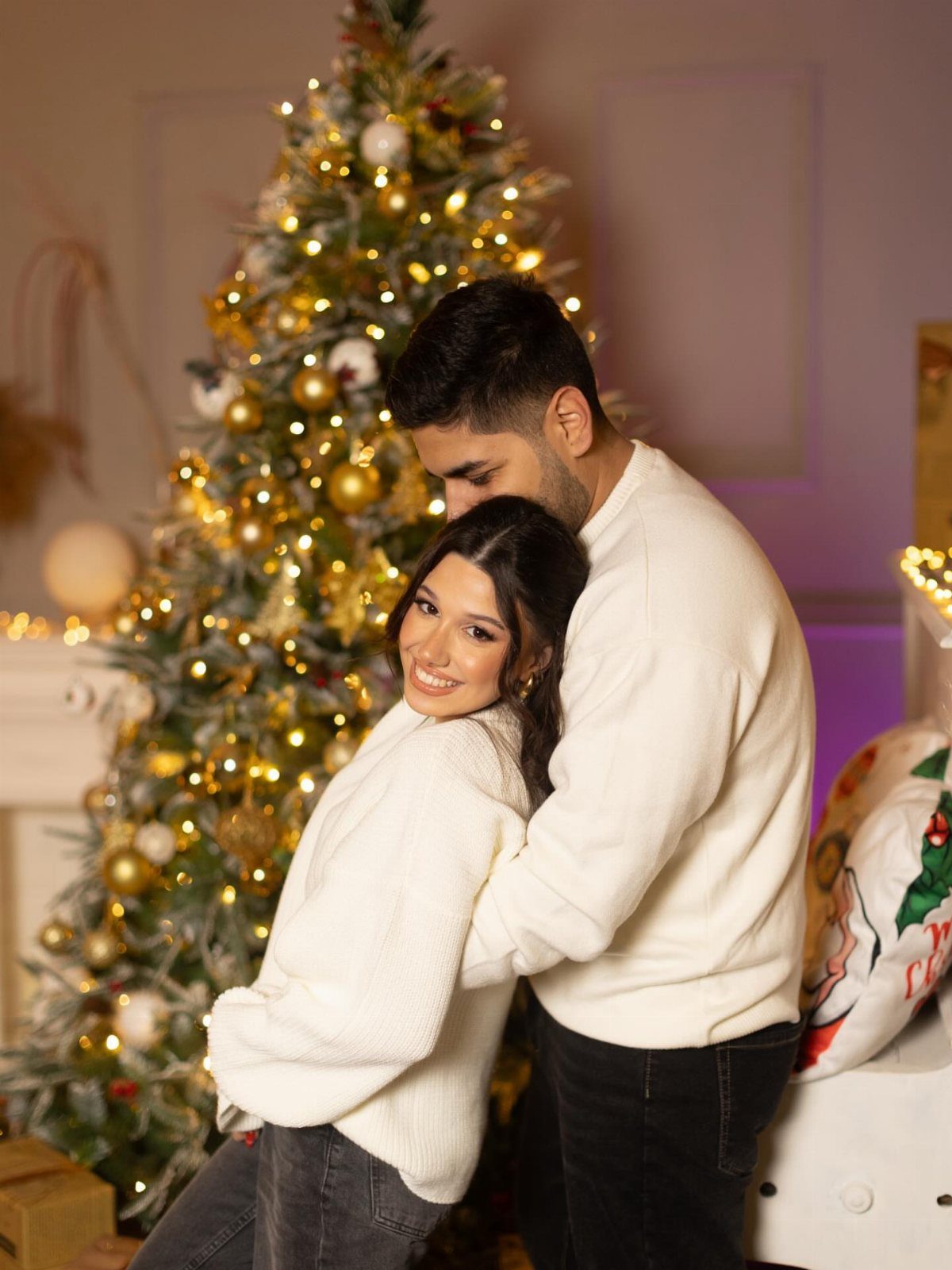 Couple embracing in white sweaters in front of a glowing Christmas tree.