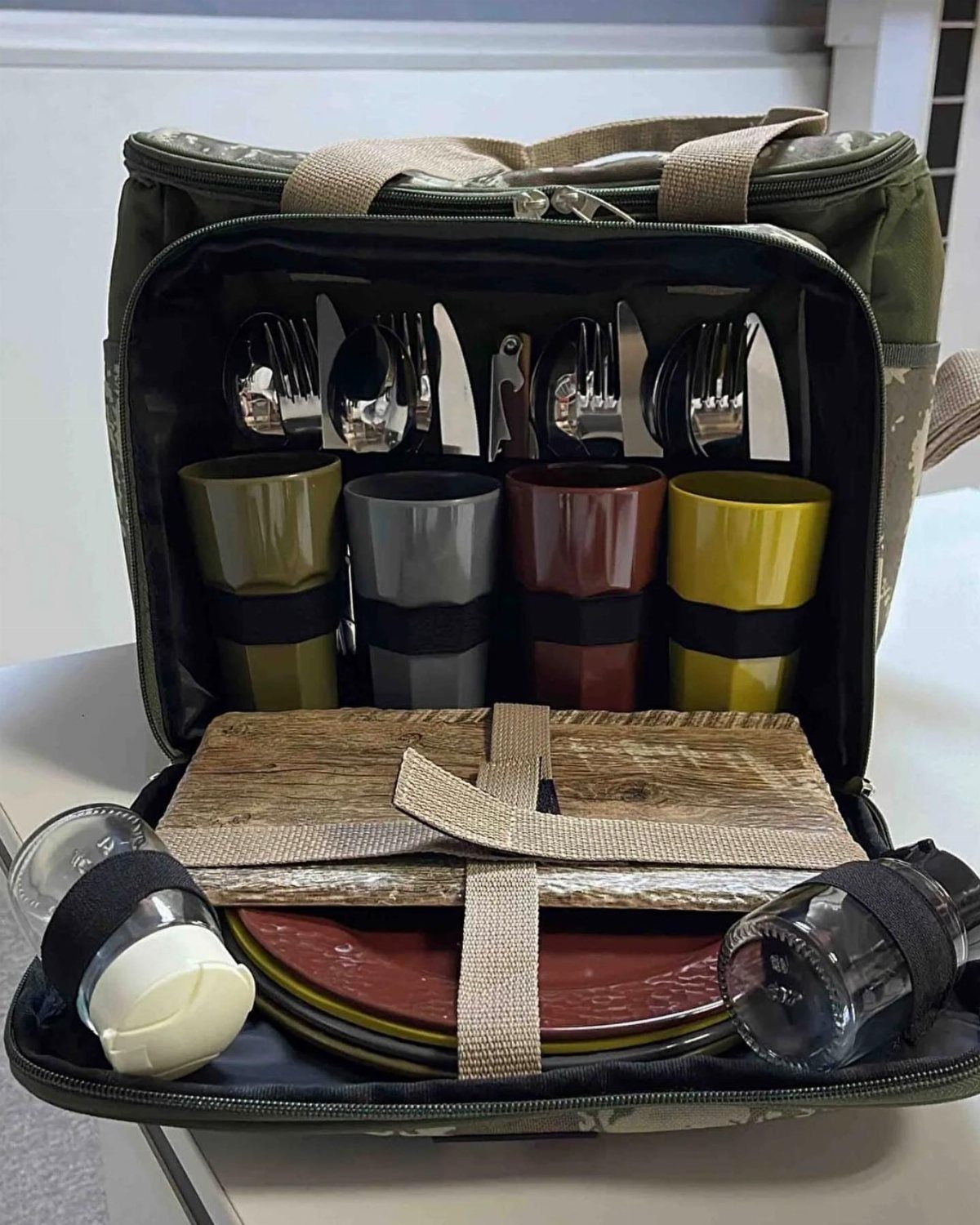 Open olive green picnic backpack with organized cups, cutlery, plates, and cutting board.