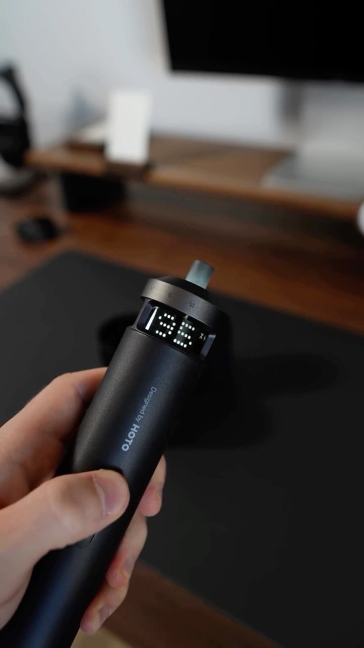 Hand holding a black electronic screwdriver with a digital LED display on a desk