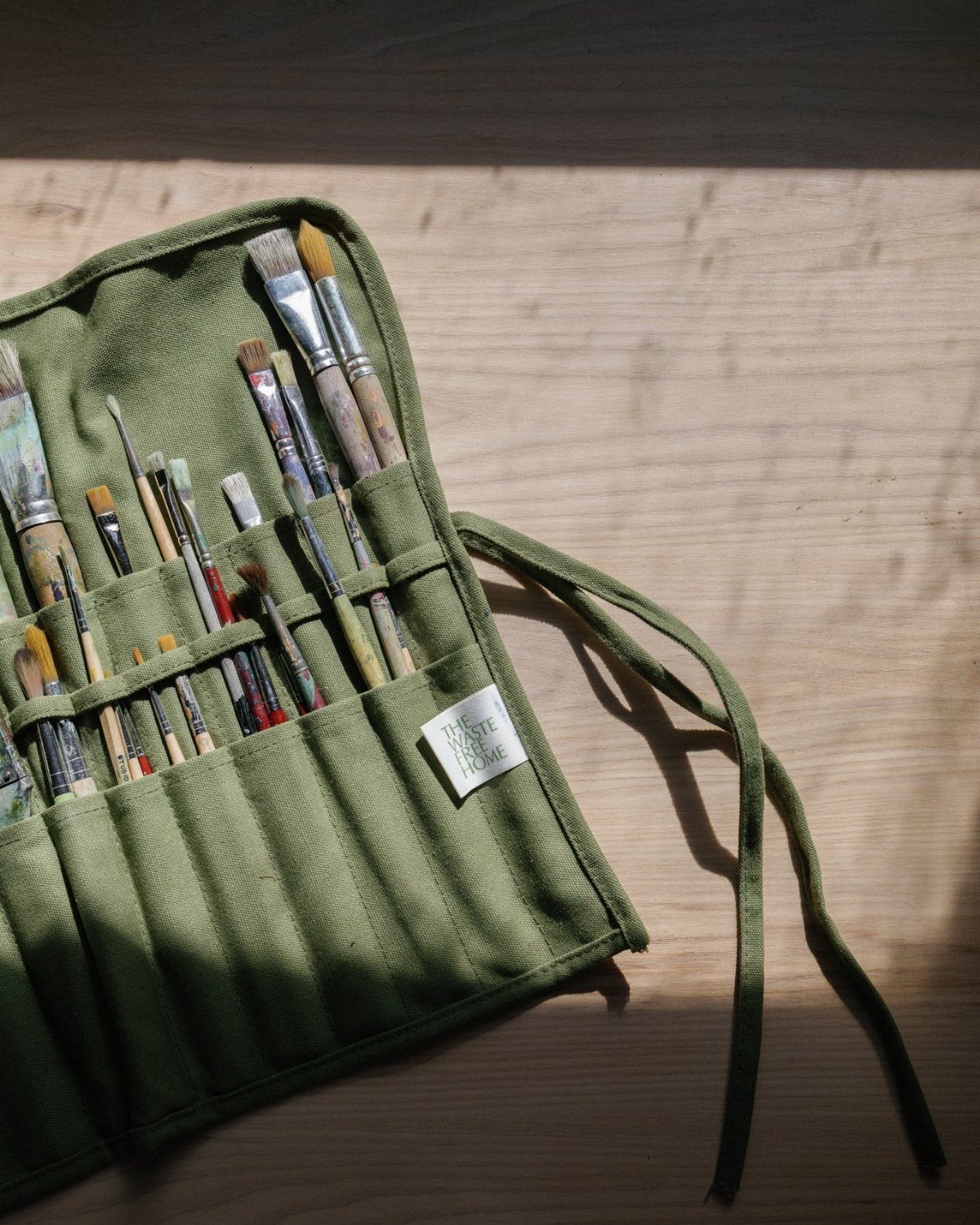 Olive green fabric brush roll holding various paintbrushes on a wooden surface.