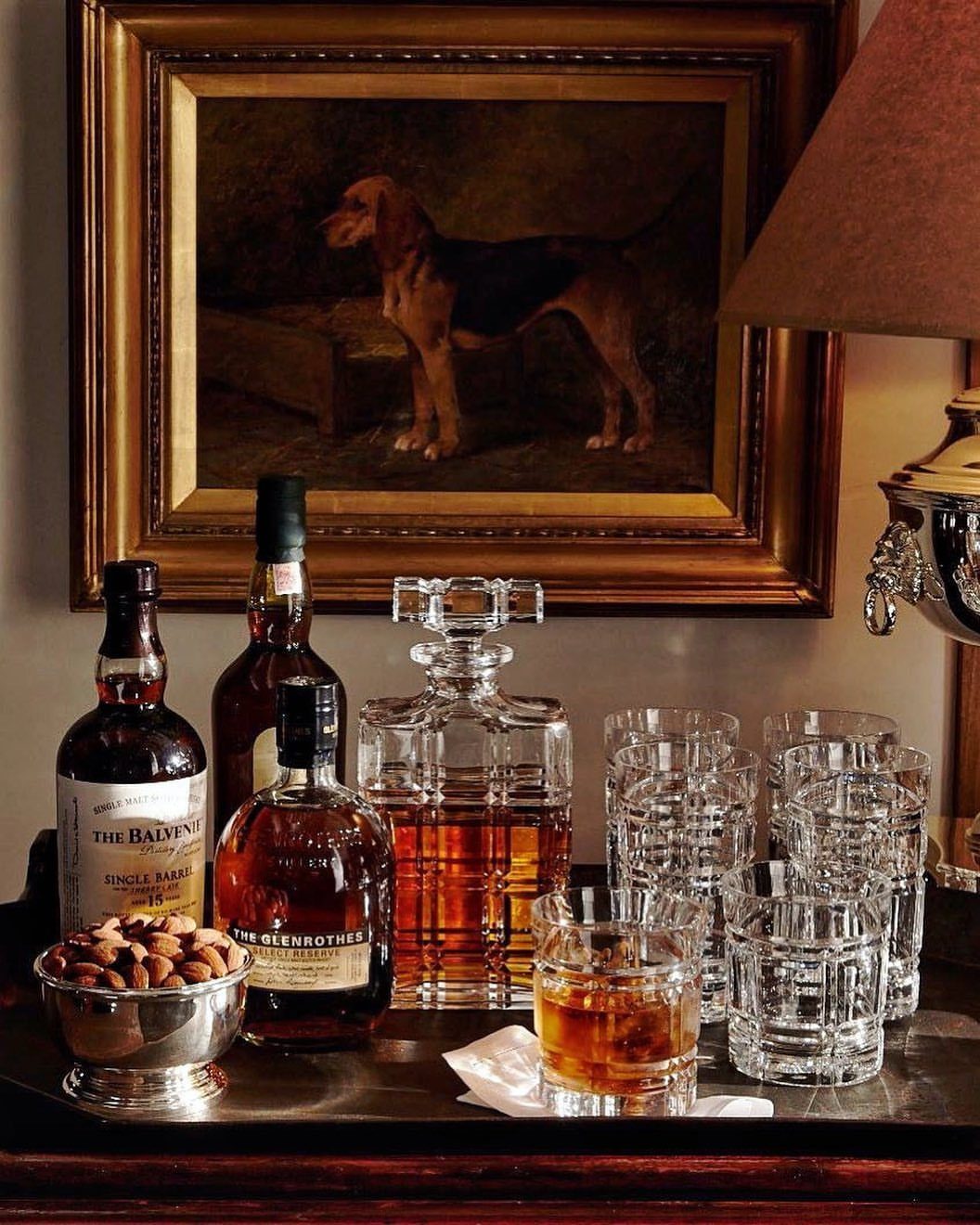 Whiskey bottles, crystal decanter, and glasses on a tray before a dog painting.