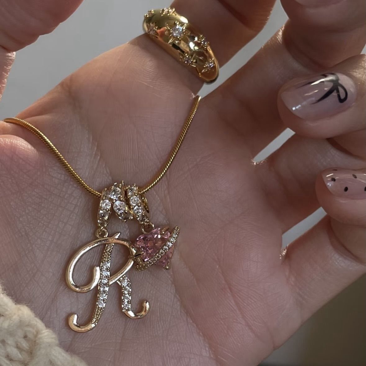 Gold chunky initial R necklace with pink heart charm held in a hand