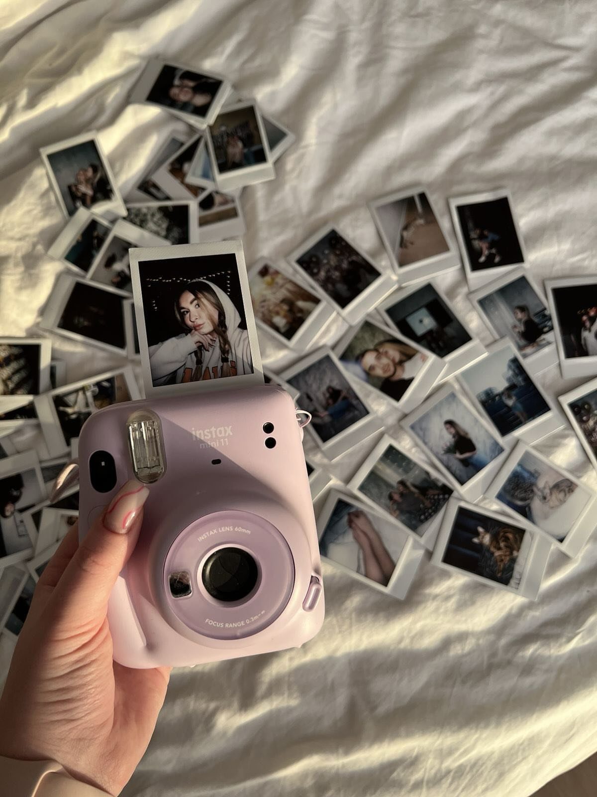 Light purple instant camera holding a printed photo over scattered polaroids on white fabric
