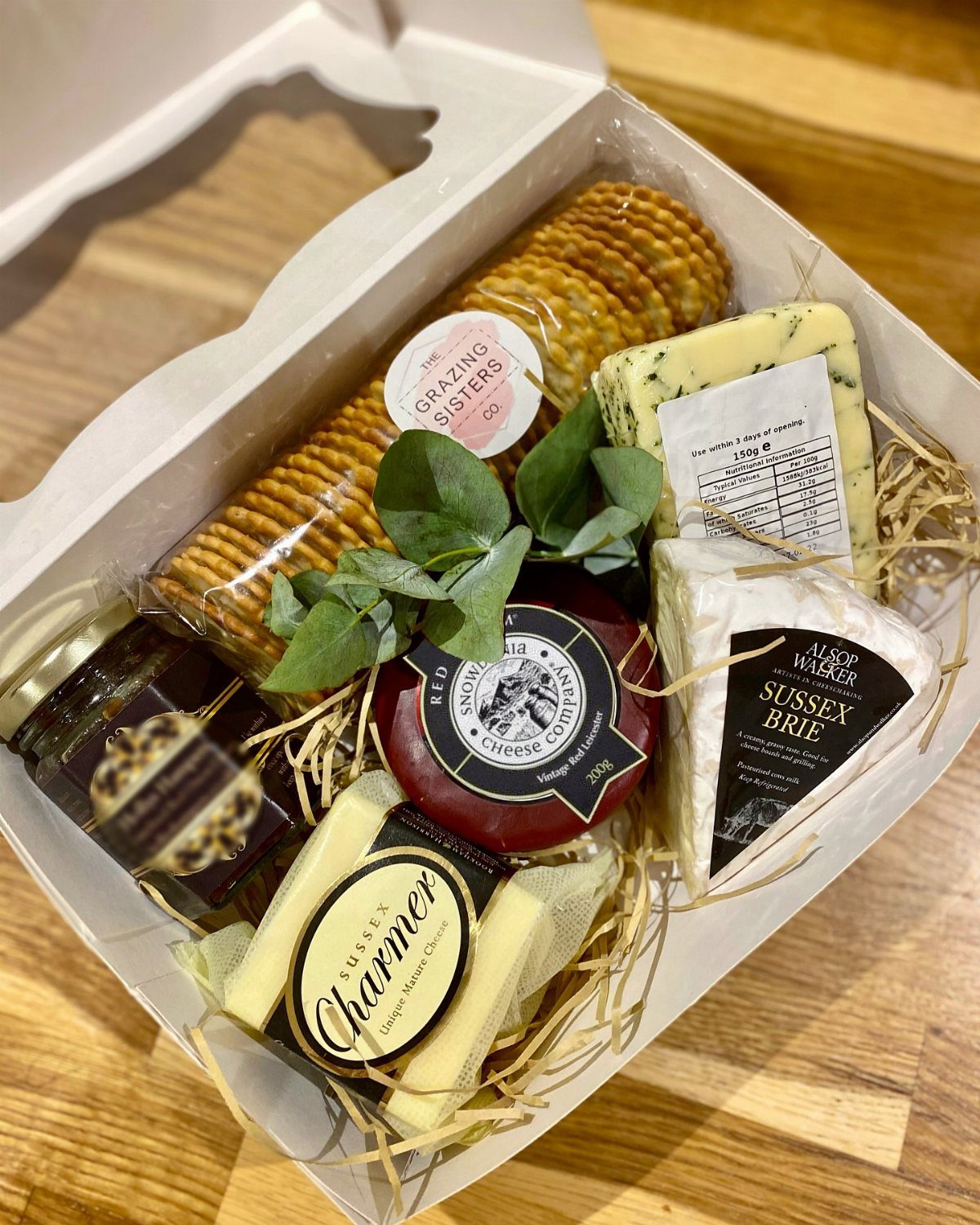 Assorted artisan cheeses, crackers, and preserves arranged in a white gift box.