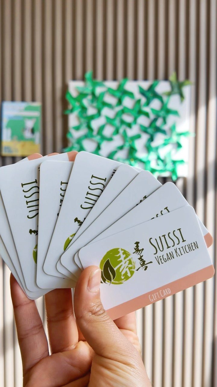 Hand holding a fan of white Suissi Vegan Kitchen gift cards against ribbed wall.
