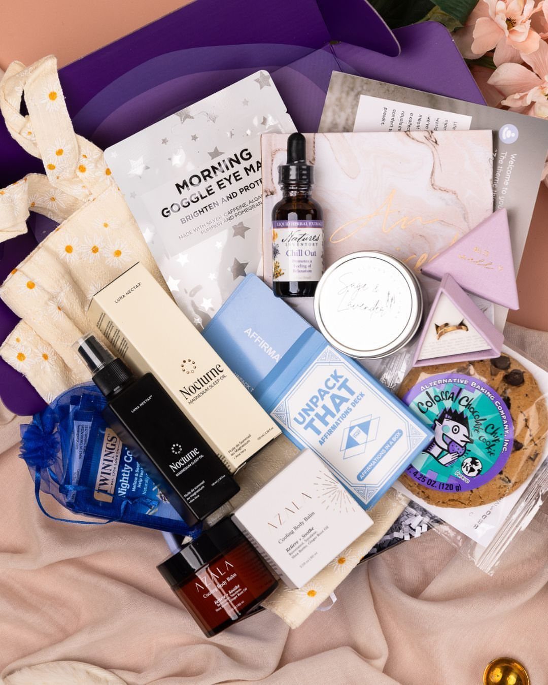 Purple gift box containing skincare products, a candle, and a chocolate chip cookie.