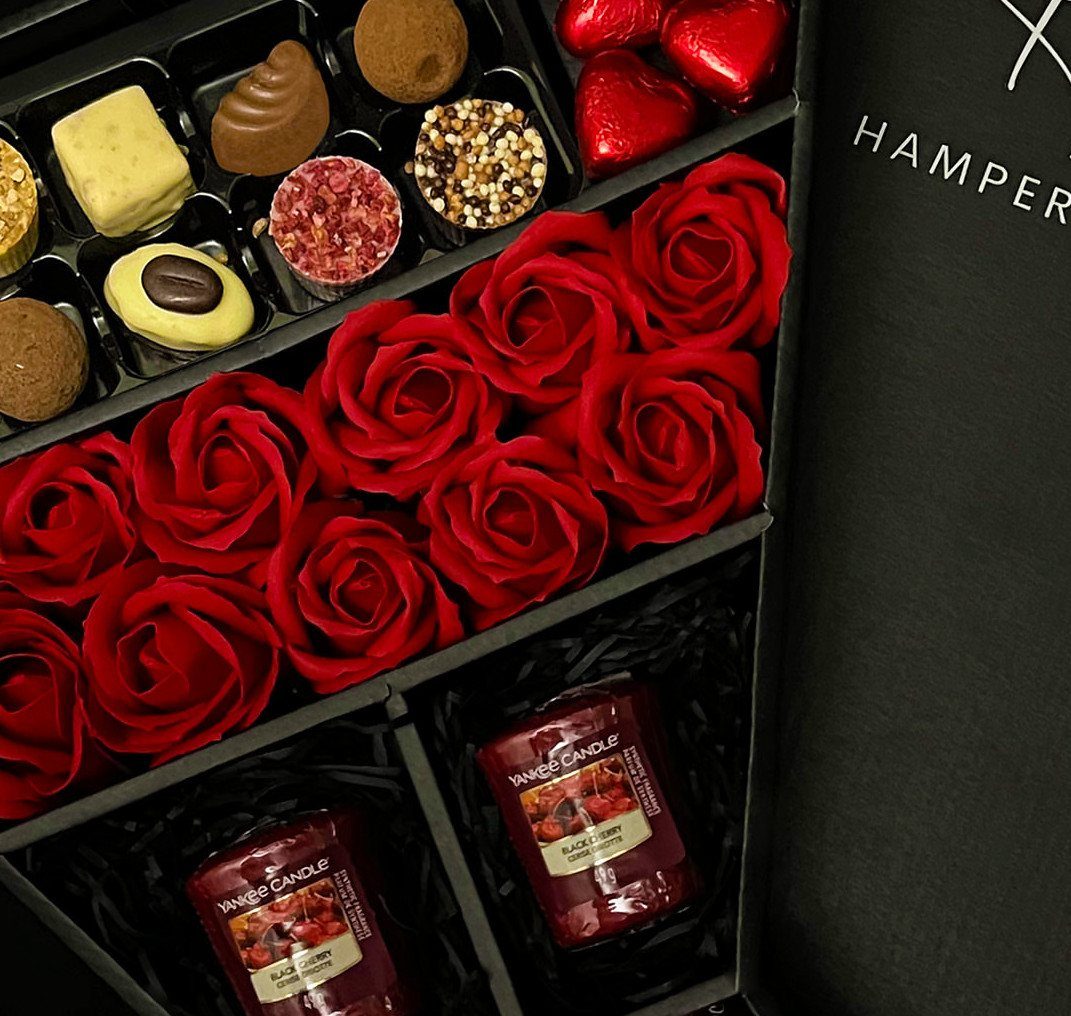 Gourmet chocolate, red roses, and scented candles arranged in a black gift hamper.