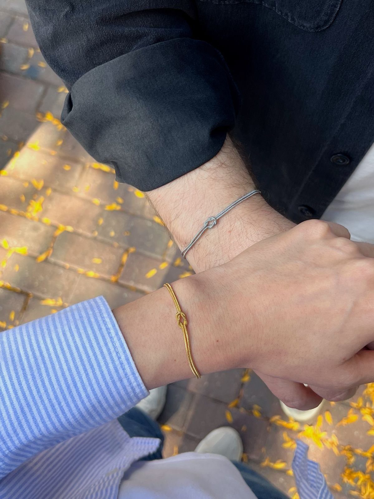Two people holding hands wearing matching braided bracelets in grey and gold colors.