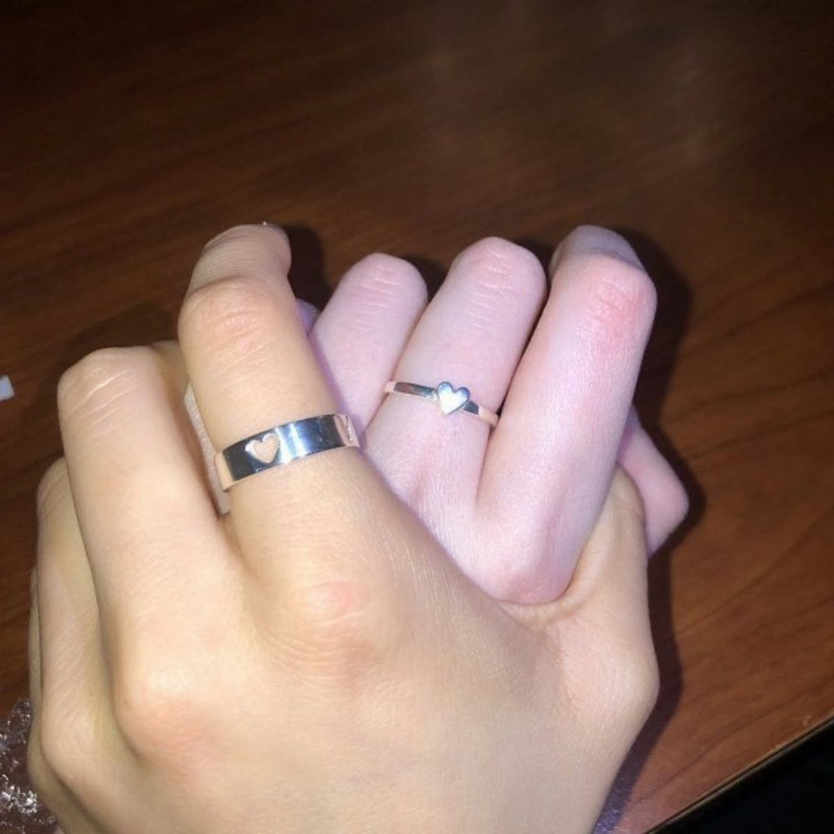 Two hands holding each other wearing matching silver heart rings on a wooden table