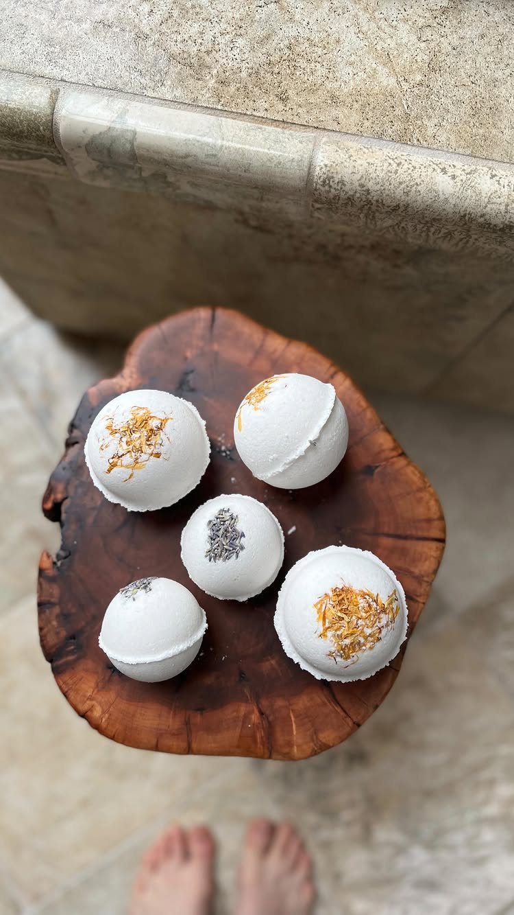 White bath bombs with dried botanicals on a rustic wooden slice by a tub
