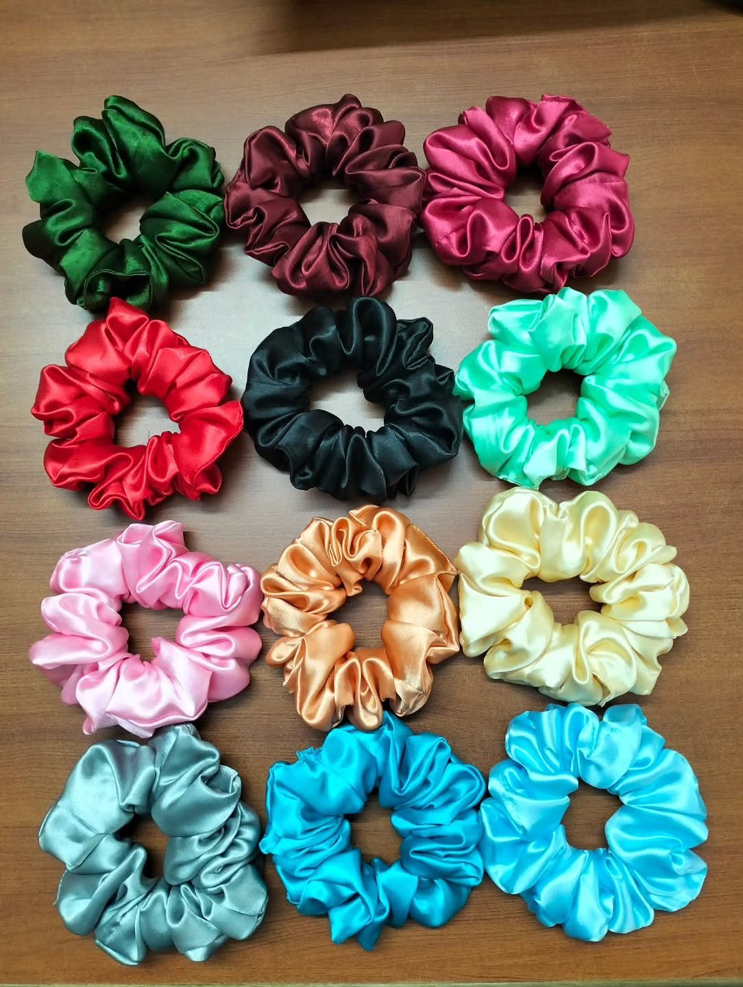 Twelve colorful satin hair scrunchies arranged in a grid on a wooden table.