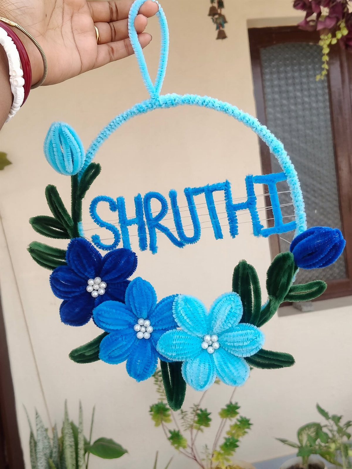 Blue circular name plate with the name SHRUTHI and fabric flowers held by hand.