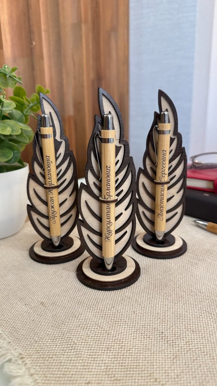 Three feather-shaped wooden pen holders with bamboo pens on a beige fabric surface.