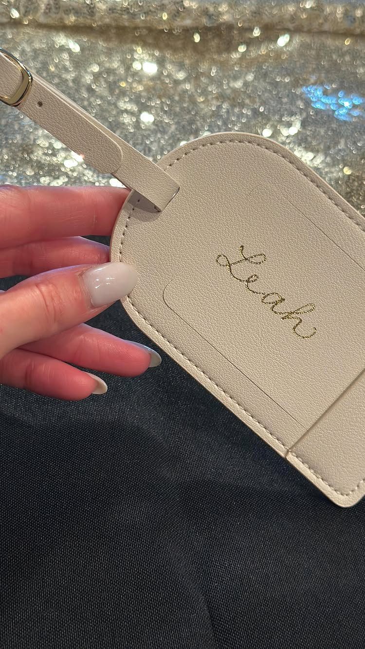 Beige personalized luggage tag with gold cursive name Leah held by a hand.