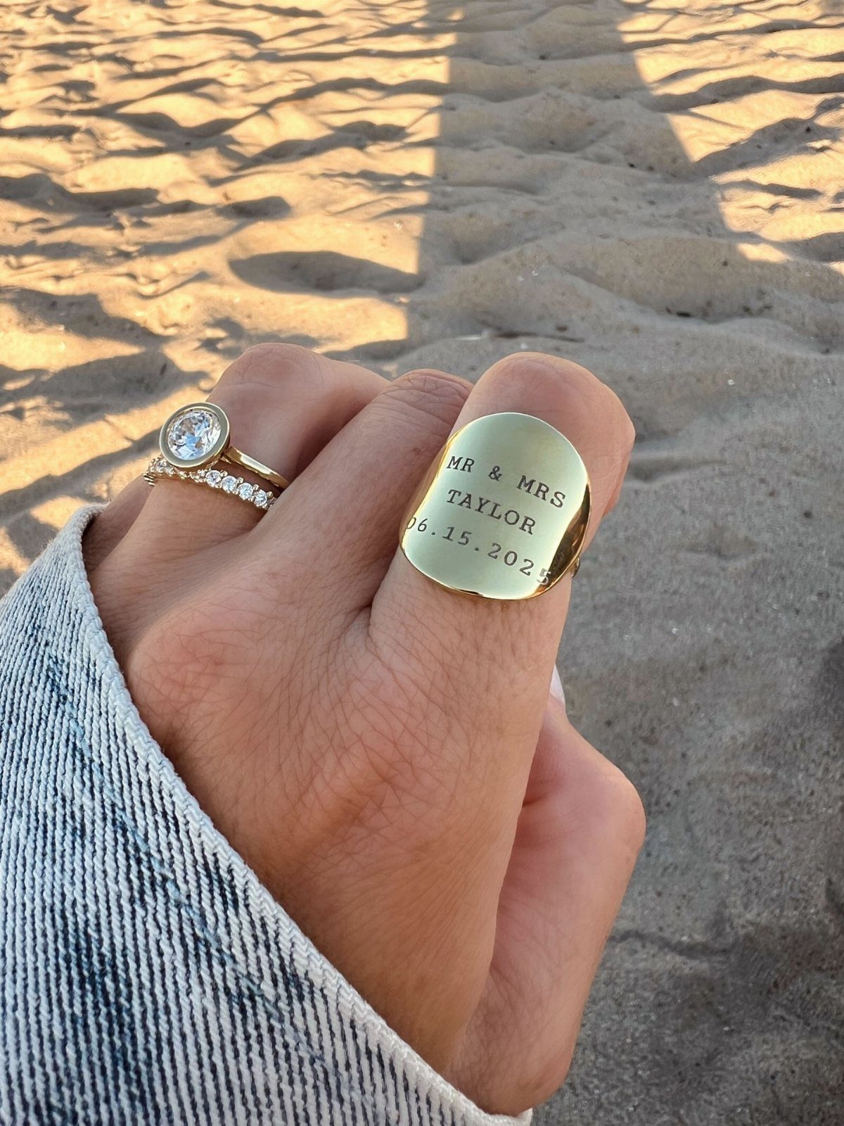 Hand wearing a diamond engagement ring and engraved gold signet ring on sand