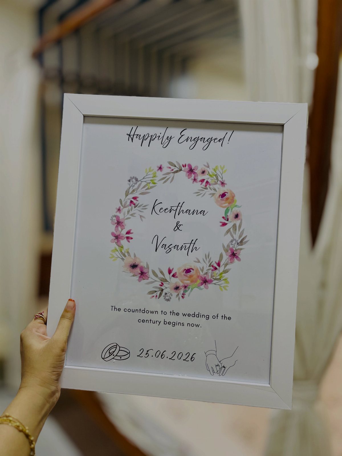Framed engagement announcement with floral wreath and names held by a hand.