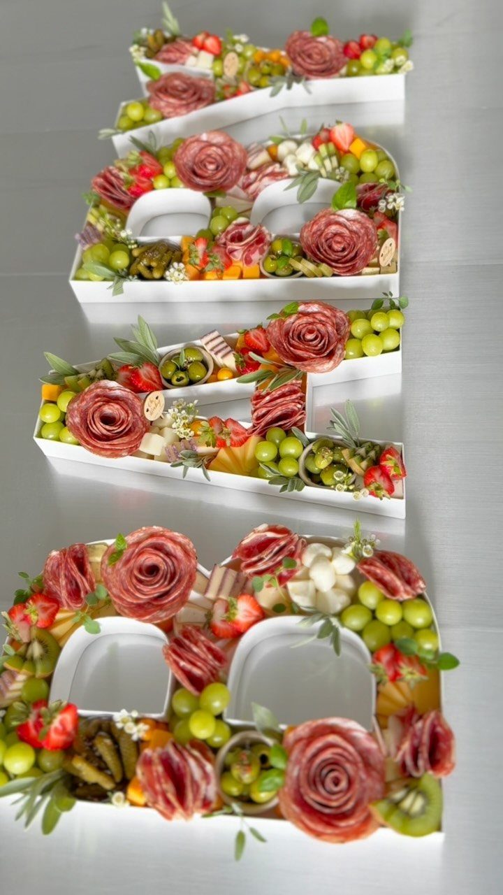 Charcuterie board shaped like the word LOVE with meat roses and fresh fruit.