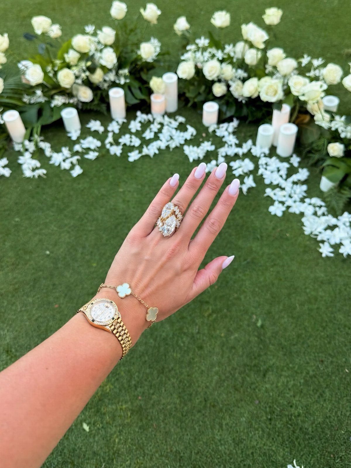 Hand wearing a large diamond engagement ring, gold watch, and bracelet outdoors.
