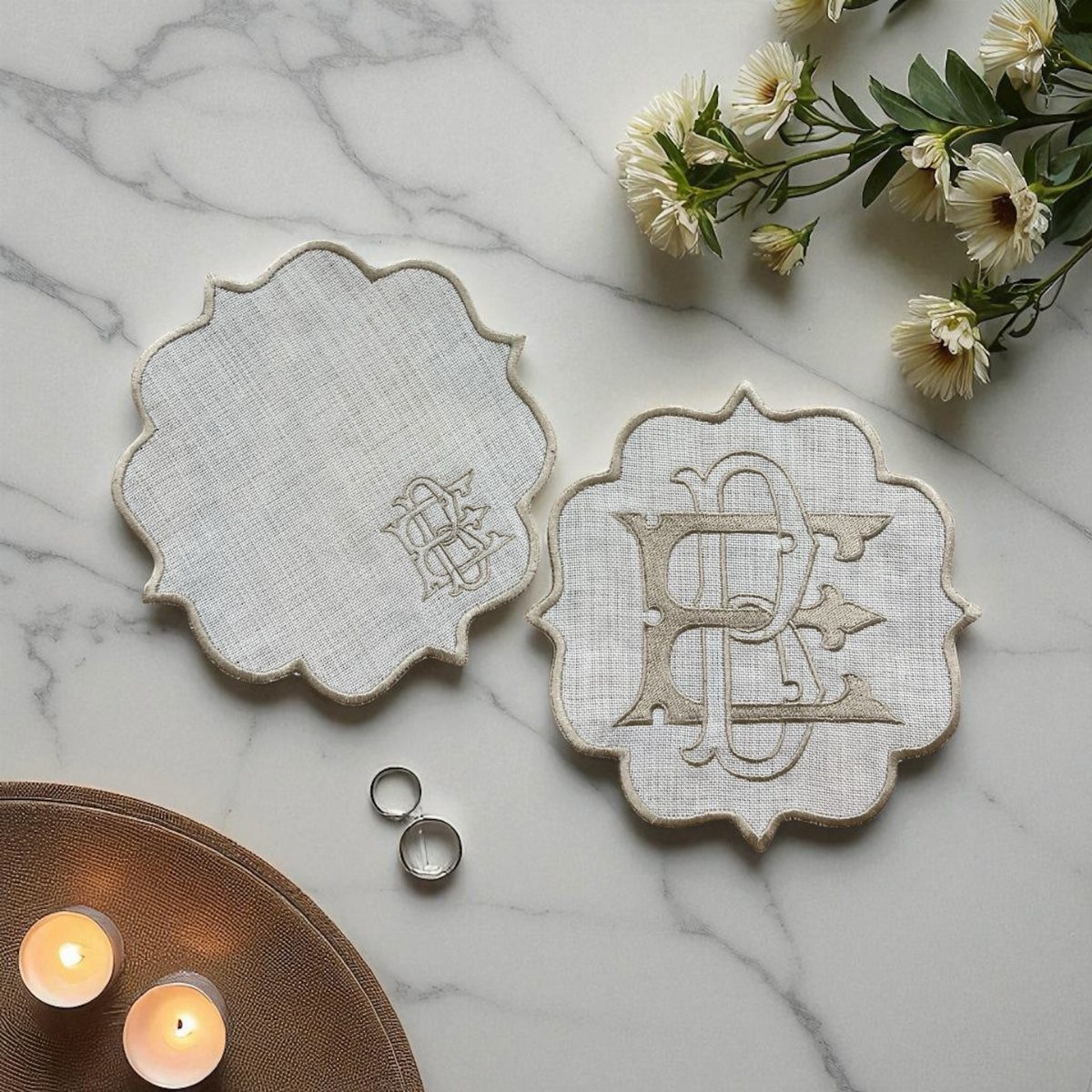 Two white embroidered monogram coasters on a marble surface with rings and flowers.