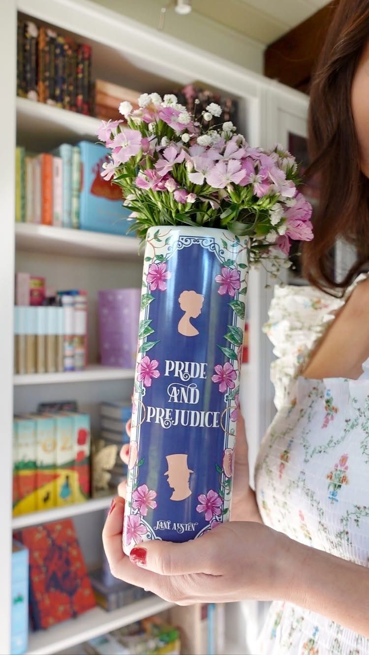 Book-shaped Pride and Prejudice vase with pink flowers held before a bookshelf.