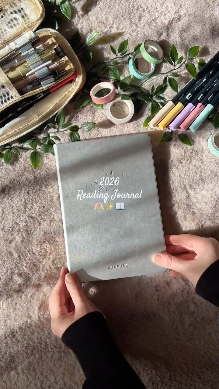 Grey reading journal held by hands surrounded by colorful markers and washi tape.