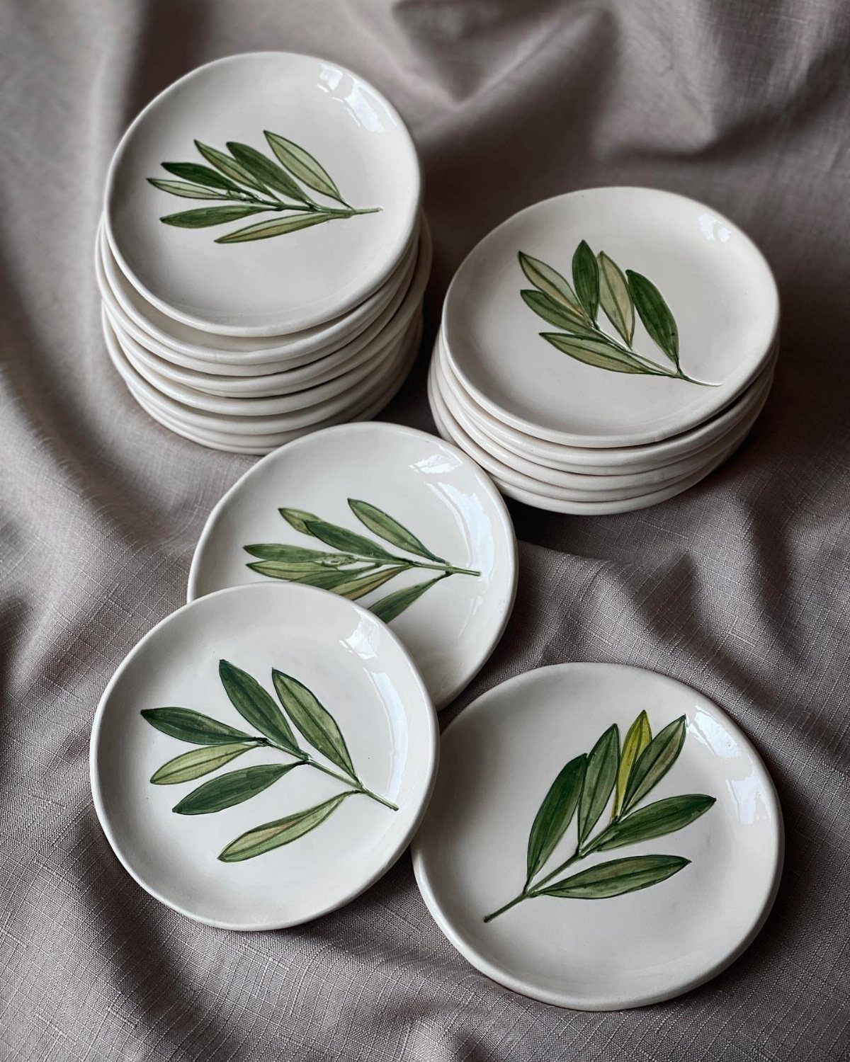 Small white ceramic dishes with hand-painted green olive branches on grey fabric.