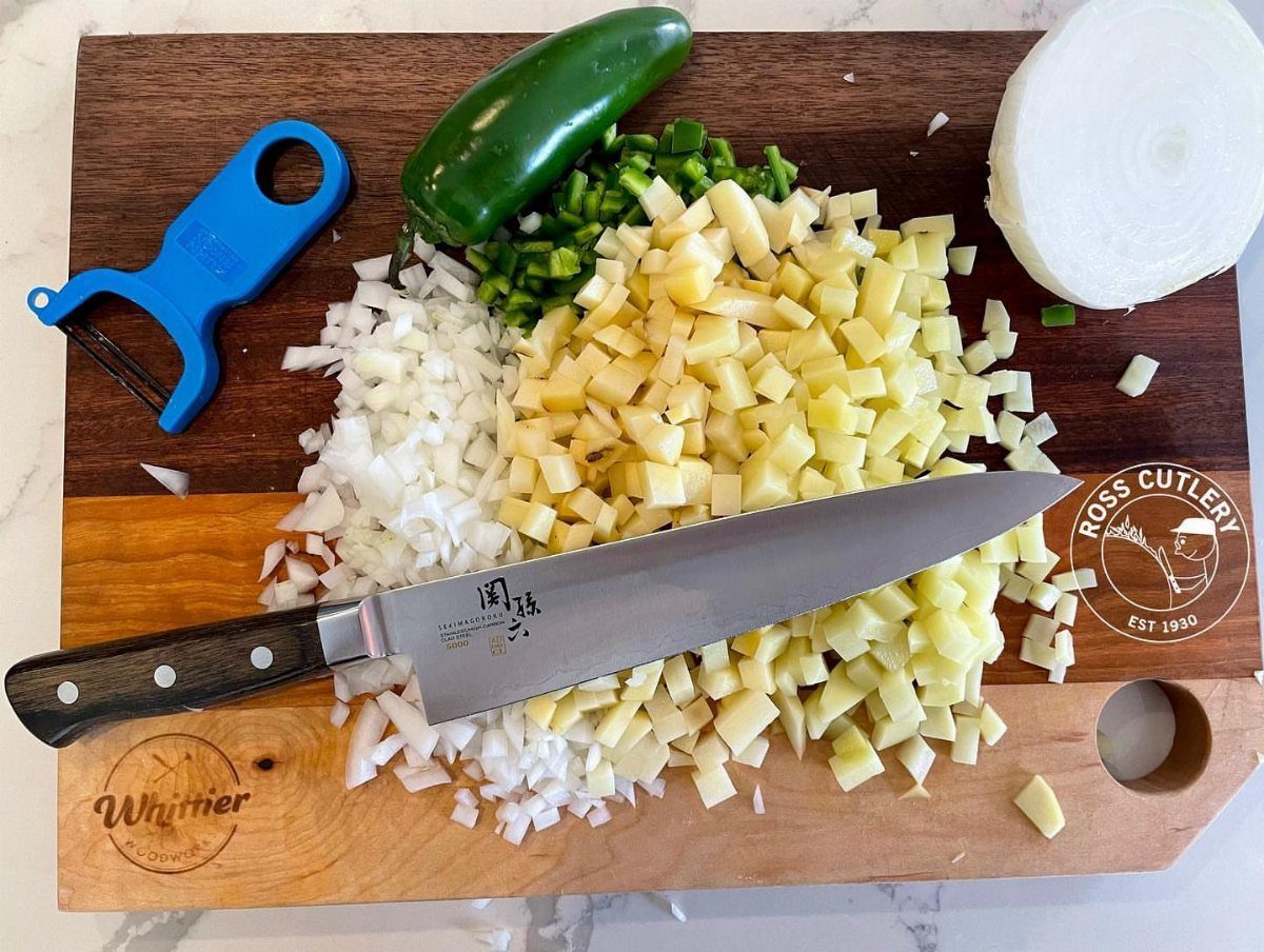 Chef's knife on a wooden board with diced potatoes, onions, and jalapeños.
