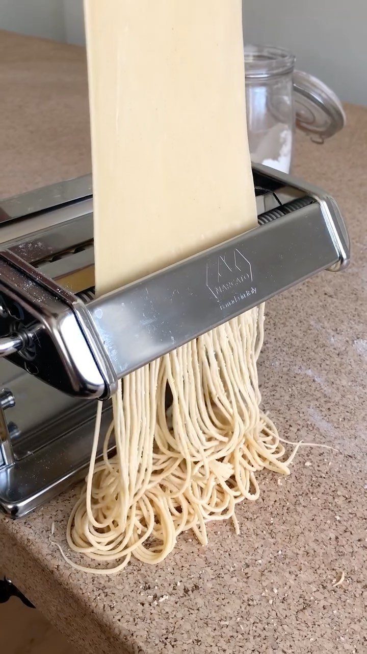 Silver pasta machine extruding fresh pale yellow noodles onto a beige countertop.