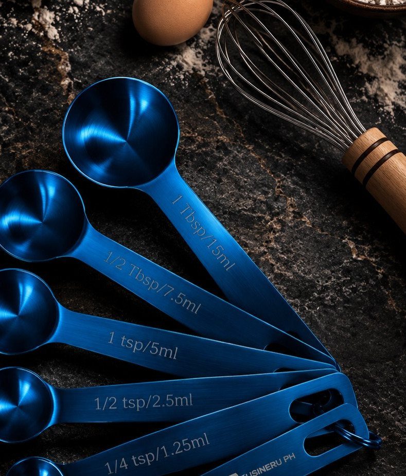 Blue metallic measuring spoons on a dark stone surface with a whisk and egg.