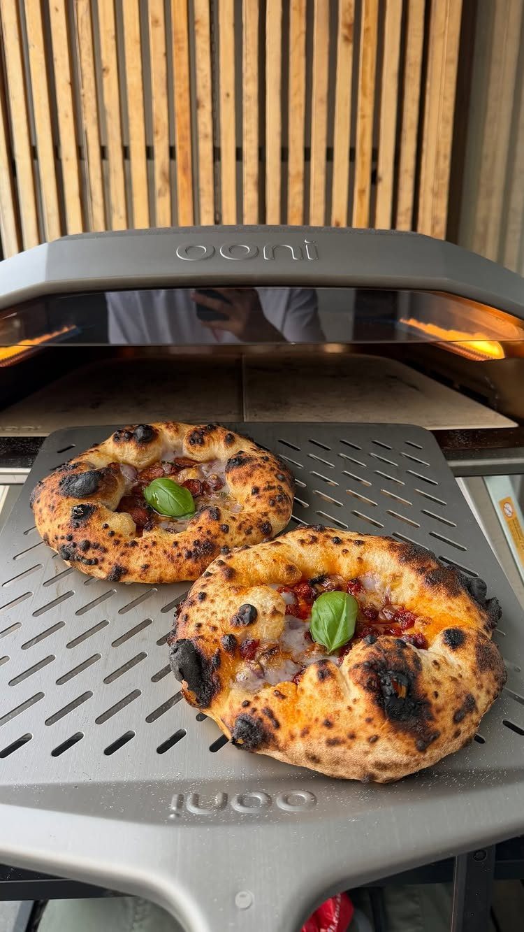 Two charred pizzas on a metal peel in front of an Ooni oven.