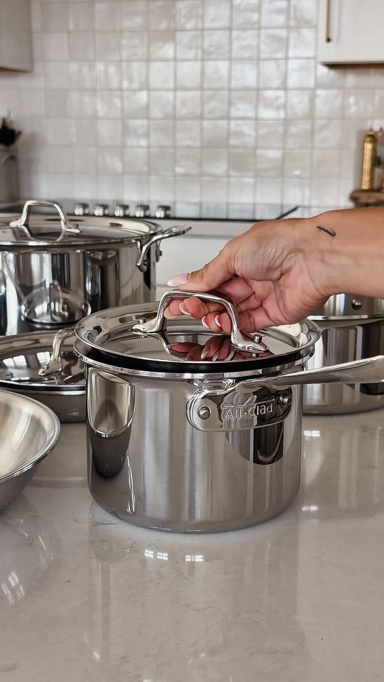 Stainless steel All-Clad pot on a white marble kitchen counter with a hand.